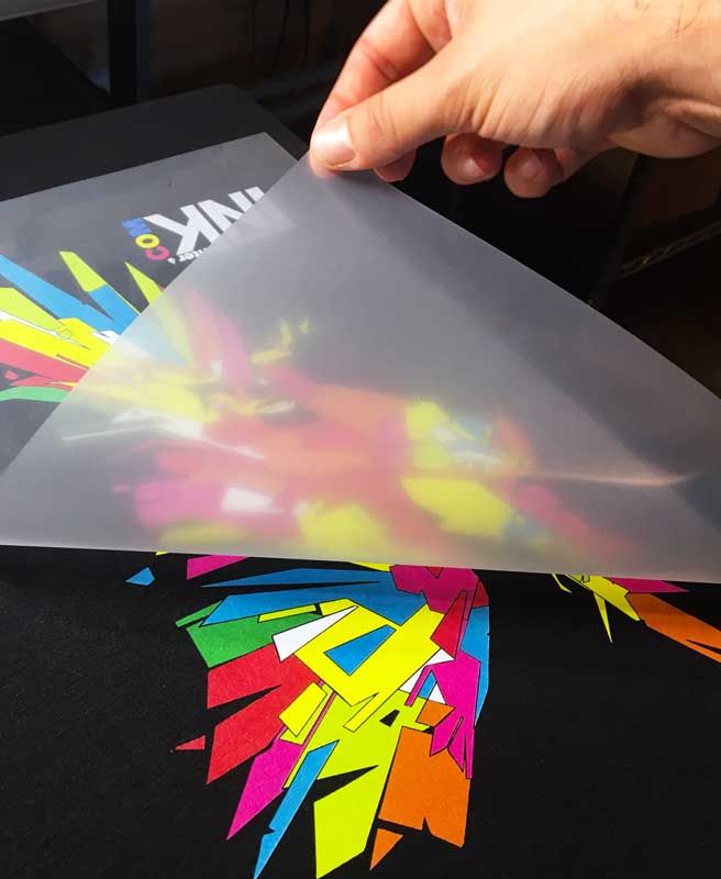 Close-up of a hand lifting a clear laser transfer sheet revealing a vibrant, geometric print design on a black fabric surface.